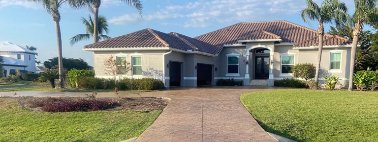 Modern single-story home with brick driveway, tile roof, dark garage doors, and palm trees on manicured lawn