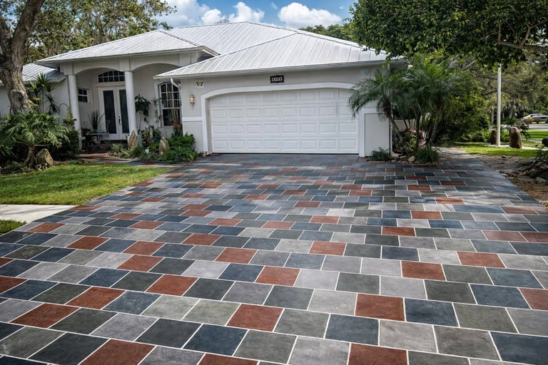 Modern single-story white house with gray metal roof and two-car garage, featuring a colorful patterned brick driveway and landscaped grounds with trees and shrubs