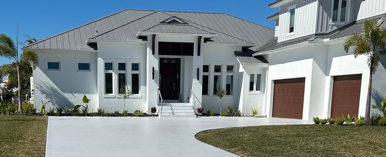 Modern white residential home with metal roof, double garage, landscaped lawn, and palm trees under clear blue sky